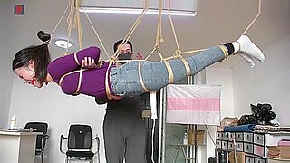 Eine hängende asiatische Mädchen erträgt BDSM-Fesseln und neckt.